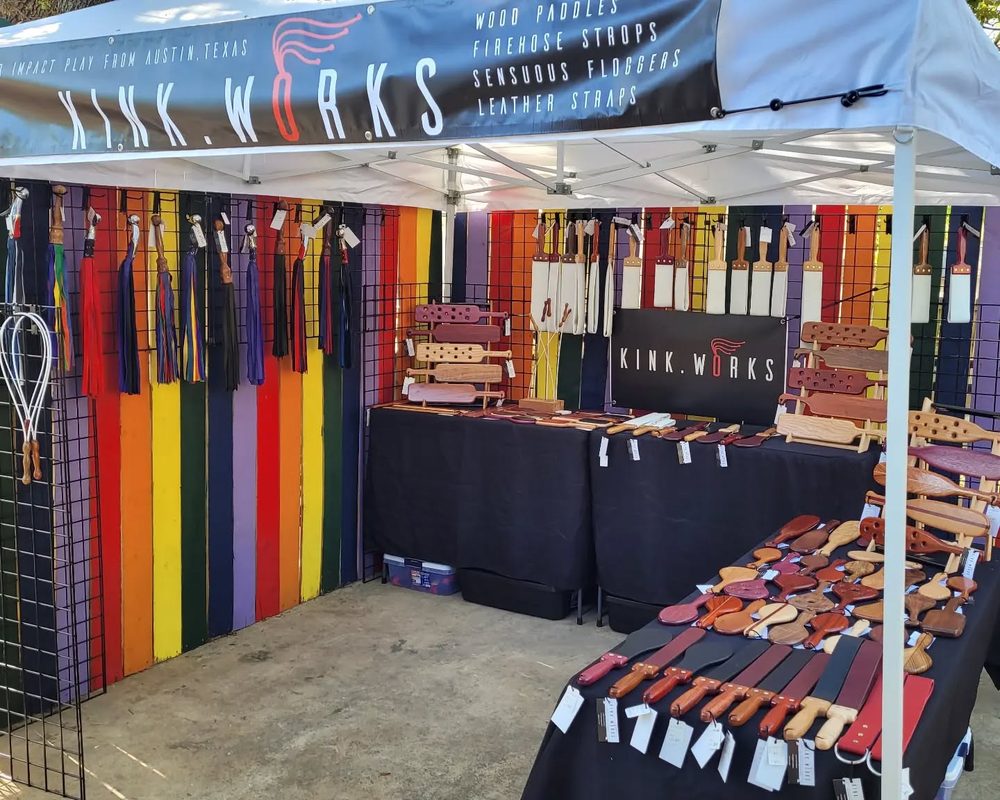 Kink.Works vending booth under a canopy with a branded banner and hanging leather