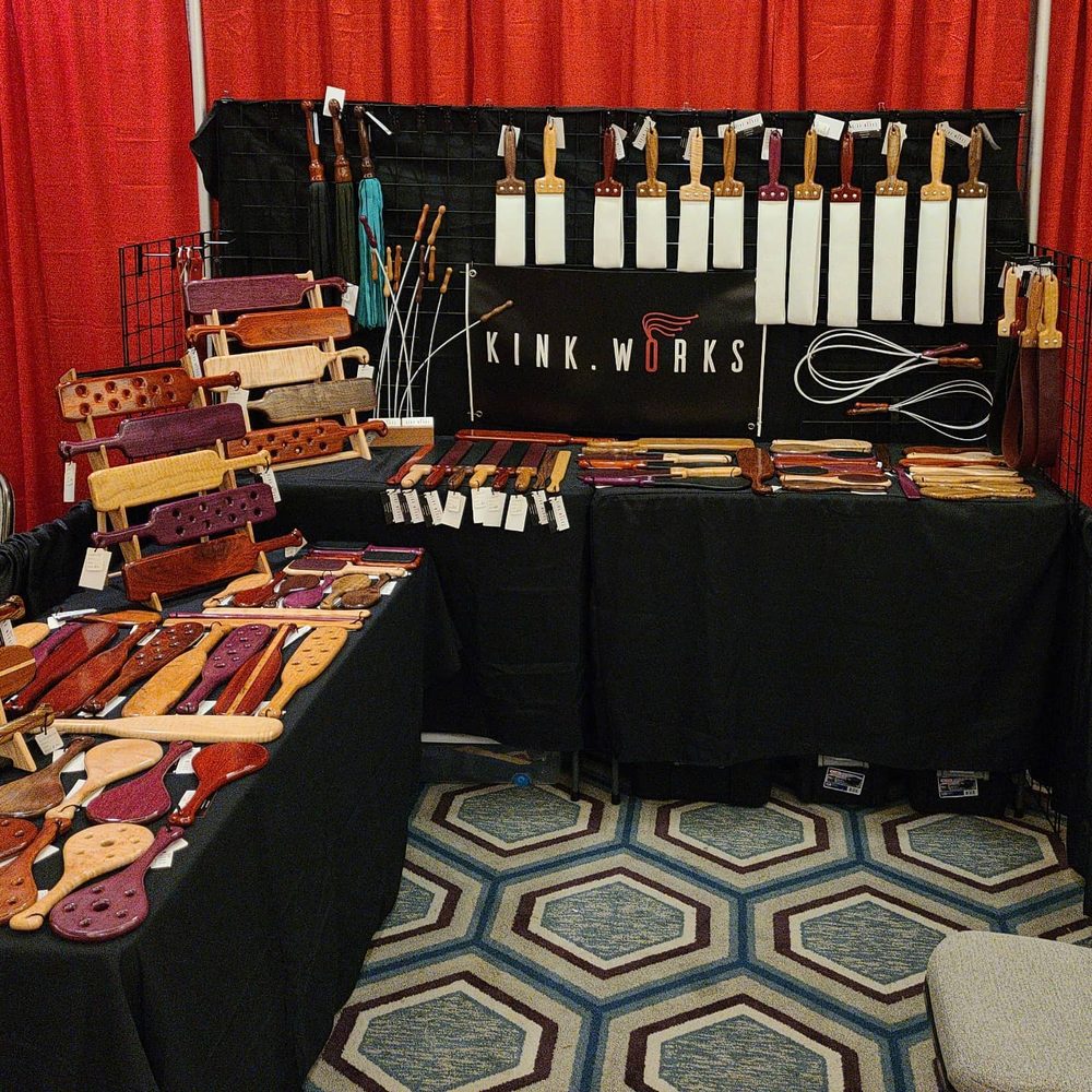 Kink.Works booth with paddle racks, hanging straps and a branded banner
